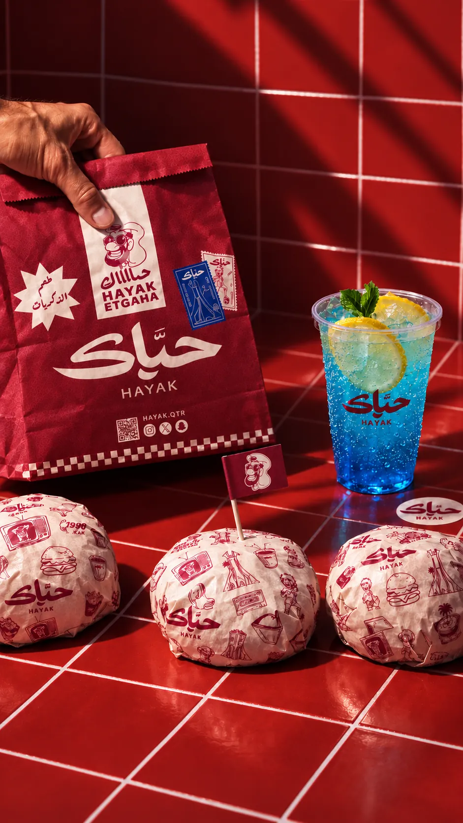 Hayak burgers wrapped in doodle paper on red tiles, with a bag and a blue soda
