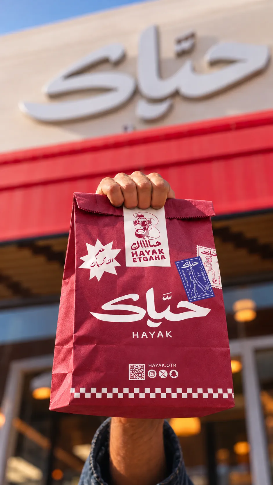 A wine-red Hayak takeaway bag held up outside a Hayak store